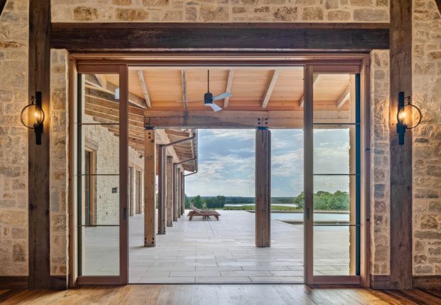 View from a stone-framed interior through large timber-framed sliding glass doors onto a broad covered terrace. The space features exposed timber beams and columns, pale stone paving, dark timber flooring, and black wall-mounted lanterns, with a calm water and landscape outlook beyond.