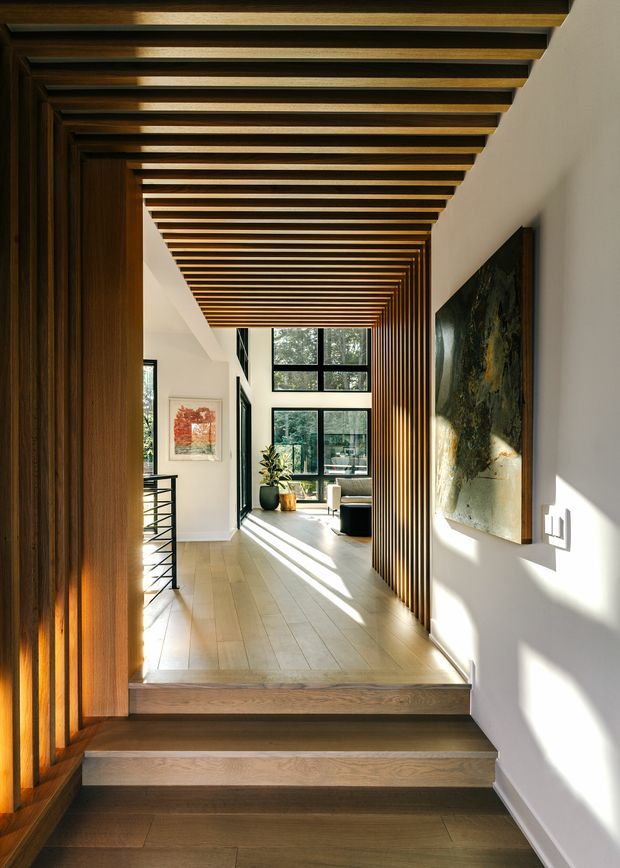 A sunlit contemporary interior hallway viewed from a short stairway, featuring a dramatic continuous ceiling and wall screen of dark timber slats. Pale timber flooring, white plaster walls, black-framed windows, a black metal balustrade, artwork, and a potted plant lead toward a bright living area. Strong linear perspective, warm timber detailing, and abundant natural light define the space.