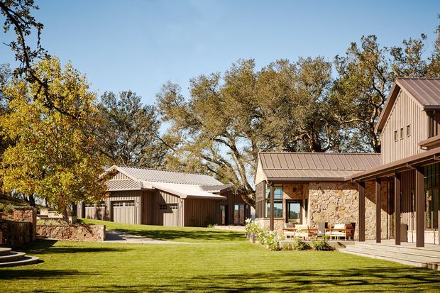 Exterior view of a low-rise residential compound arranged around a broad manicured lawn. The buildings feature vertical timber siding, pitched standing-seam metal roofs, deep roof overhangs, covered verandas, large glazed openings, and a prominent natural stone wall section. Mature deciduous trees and planted beds frame the courtyard-like setting in warm autumn light.