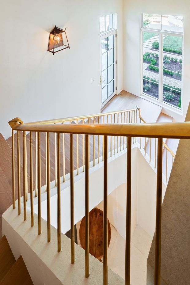 Bright interior view of a multi-level stairway and landing, looking down through a sculptural stairwell. The staircase has warm timber handrails and slender vertical metal balusters, pale painted walls, light timber flooring, and a geometric wall lantern. Tall divided-pane windows and a glazed door bring in abundant daylight, with a landscaped garden visible outside.