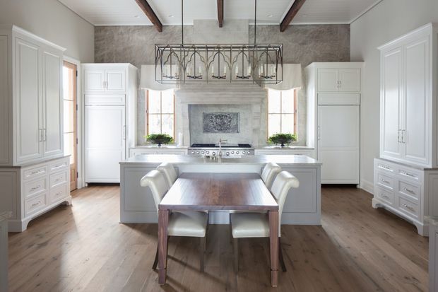Bright, spacious kitchen with a central island and integrated cooktop, surrounded by extensive white paneled cabinetry. A timber dining table with upholstered chairs sits in the foreground. The room features wide-plank timber flooring, a large stone-look rangehood and backsplash wall, exposed dark timber ceiling beams, and a substantial rectangular pendant light over the island. Tall windows bring in abundant natural light.