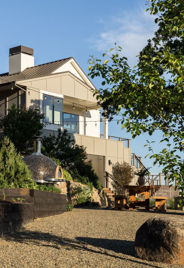 Exterior view of a contemporary multi-level home on a landscaped sloping site, featuring a steep gable roof, light panelled facade, broad outdoor terrace with glass balustrades, exterior stairs, festoon lighting, gravel paving, timber retaining walls, and a distinctive domed outdoor fireplace or oven in the foreground. Mature planting partially screens the building.