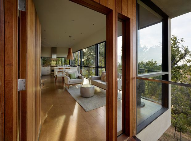 View through an open timber-framed entry door into a bright, modern open-plan living area with a visible kitchen at the rear. The interior has extensive black-framed glazing, warm timber wall and door surfaces, pale flooring, neutral upholstered seating, and a contemporary pendant light. A glazed balcony or terrace and leafy exterior views are visible beyond the living room.