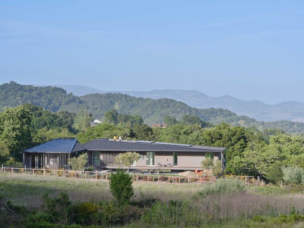 A contemporary single-storey house set in a hilly, wooded landscape. The long low-profile building has dark metal pitched roofs, extensive glazing, and a restrained neutral facade, with a deck or terrace running along the front. The surrounding rural garden and layered mountain backdrop are clearly visible.