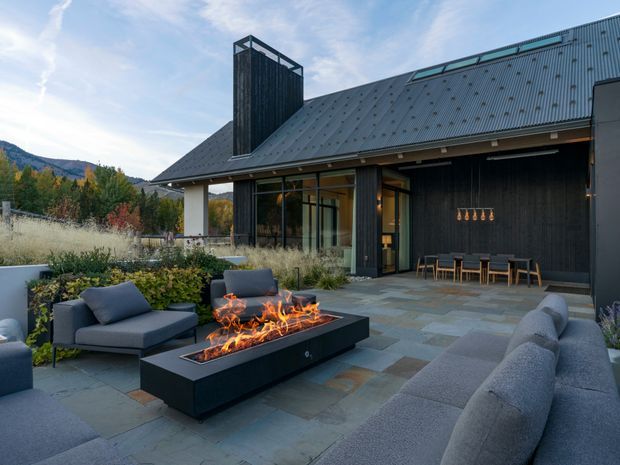 A contemporary mountain-side home viewed from a landscaped outdoor entertaining area. The dark timber-clad building has a steep corrugated metal roof, expansive glazing, and a tall enclosed chimney or flue feature. A paved terrace includes a long linear outdoor fire table, built-in and freestanding grey seating, and an outdoor dining setting. Autumn planting and mountainous terrain are visible beyond the property.
