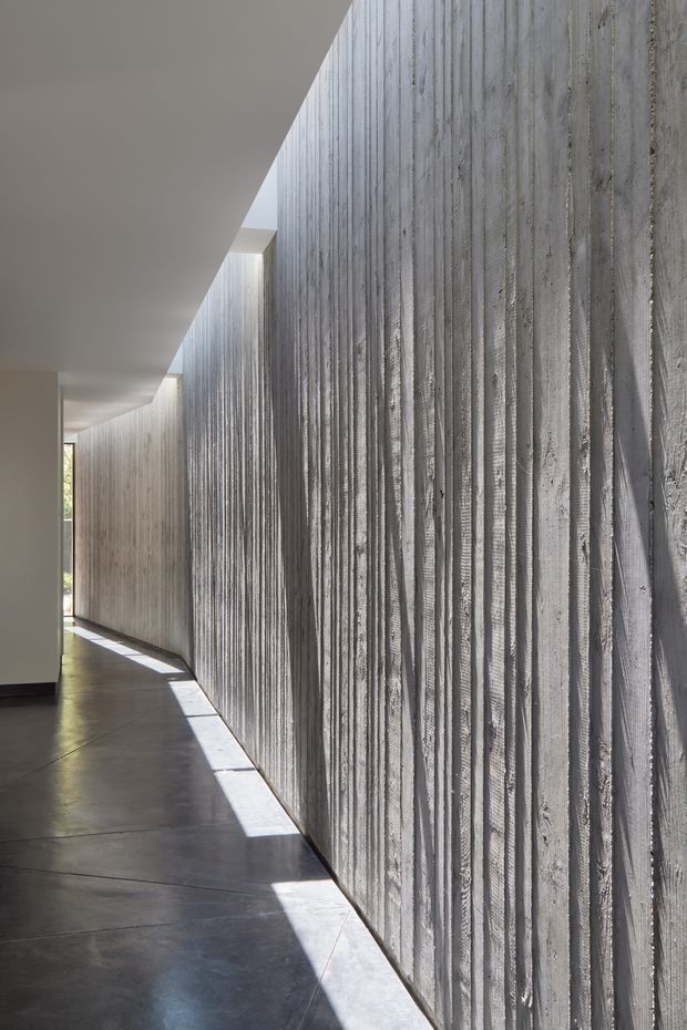 A narrow, minimalist interior corridor dominated by a long, vertically textured grey concrete feature wall. Smooth pale ceilings and walls contrast with the dark polished concrete floor, while strong linear daylight enters from openings along the side and creates bright geometric bands across the floor. The image emphasizes material texture, shadow, and architectural geometry.