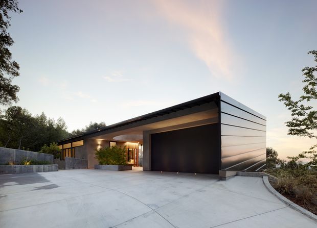 Exterior view of a low-slung contemporary residence at dusk, featuring a broad flat or very low-pitched roof, dark horizontal metal cladding, a large integrated garage door, concrete driveway and entry court, and warm-lit glazing recessed beneath a deep overhang. The home is set among trees on a slightly elevated site.