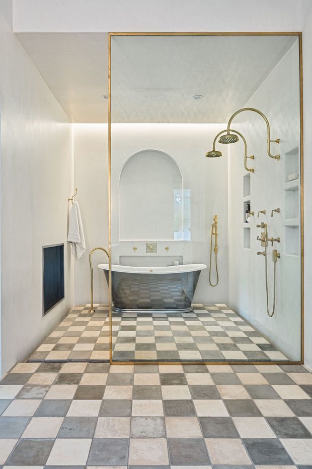 A refined bathroom with a freestanding bathtub, arched wall niche, extensive built-in shower hardware, and recessed shelving. A large frameless glass shower enclosure with a slim brass-toned frame dominates the view. The room features white plastered walls, integrated lighting, and a checkerboard floor of alternating pale stone and grey tiles, creating a classic yet luxurious appearance.