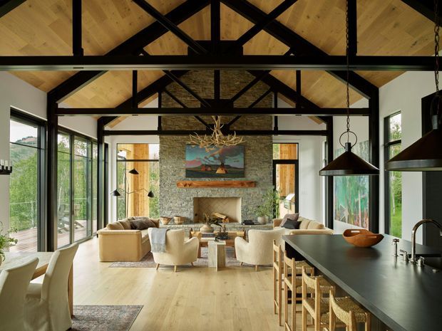 A spacious open-plan living, dining and kitchen interior with exposed black timber roof trusses, warm timber ceiling and flooring, expansive dark-framed glazing, and a prominent natural-stone fireplace wall. The room includes a large kitchen island with stools, dining seating, sofas and lounge chairs, plus sculptural pendant lighting and rustic-natural decorative elements. The design combines contemporary open planning with lodge-like, organic materials and structure.