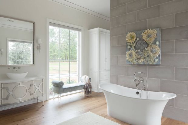 Bright, elegant bathroom with a freestanding white bathtub positioned against a grey rectangular tiled feature wall. The room includes a white double-vanity with curved decorative detailing, a vessel basin, large framed mirror, tall white storage cabinet, and a window-side upholstered bench. Warm timber flooring, white walls, and botanical wall art create a refined traditional interior.