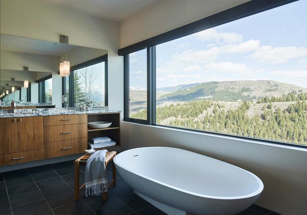 A contemporary bathroom with a freestanding white bathtub positioned beside expansive corner windows overlooking a wooded mountainous landscape. The room features dark large-format floor tiles, warm vertically fluted timber vanity cabinetry, a dark stone or granite benchtop with integrated basin, frameless mirror, black window frames, and a simple pendant light. The space is bright, refined, and strongly connected to the surrounding view.
