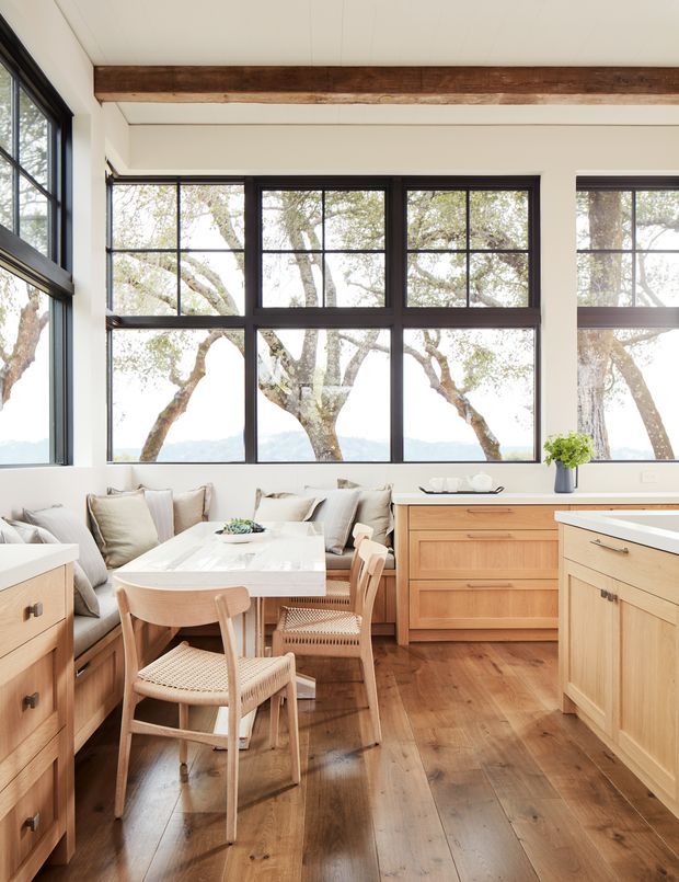A bright, compact kitchen-dining area with extensive black-framed grid windows overlooking trees. The room features light timber cabinetry, a white stone benchtop and dining table, a built-in upholstered banquette with neutral cushions, woven dining chairs, warm timber flooring, and an exposed dark timber ceiling beam. The space has a relaxed, natural, contemporary character.
