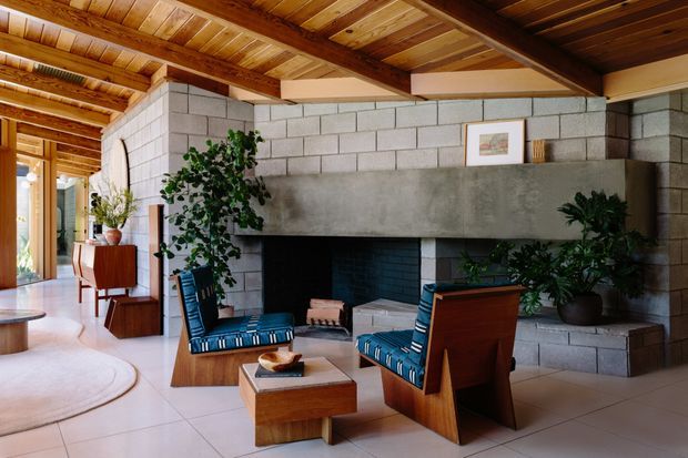A warm, mid-century-inspired living room with a large concrete-block fireplace and broad concrete mantel, exposed timber ceiling and beams, timber-framed glazing, and distinctive low wooden lounge chairs upholstered in blue striped fabric. Potted plants and a small timber coffee table complement the earthy, natural material palette.