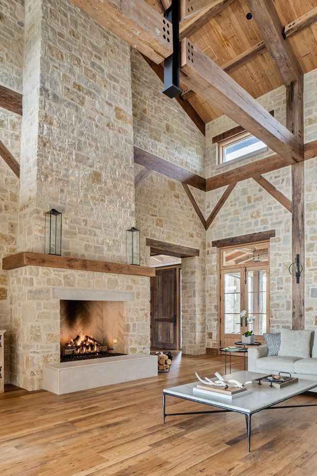 A grand rustic living room with exposed timber post-and-beam construction, a very high vaulted ceiling, and pale natural stone masonry walls. A large open fireplace with a simple stone surround anchors the room, while timber flooring, tall glazed doors, clerestory windows, and contemporary seating create a warm, refined lodge-like interior.