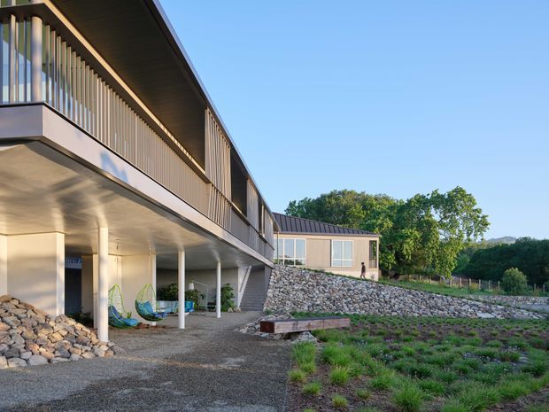 Exterior view of a contemporary elevated building with a long upper-level balcony screened by vertical timber battens, slim white columns, and a sheltered lower-level outdoor area. The building sits on a landscaped sloping site with extensive exposed stone retaining walls and gravel paving, with a low-pitched roof and large glazed openings visible on the adjoining wing.