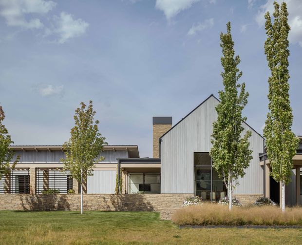 Exterior view of a contemporary single-storey residence with a prominent steep gable volume, light vertical weatherboard cladding, low stone masonry walls, extensive glazing, and a dark flat-roofed connecting wing. Tall slender trees and ornamental grasses frame the landscaped frontage.