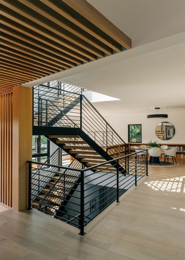 A bright, open-plan interior centered on a sculptural multi-level staircase with black metal horizontal railings and timber treads. The staircase connects to a mezzanine or upper landing beside large floor-to-ceiling glazing. Warm timber slatted ceiling beams and vertical timber wall elements contrast with white ceilings, pale timber flooring, and black metal detailing. A dining area with a table, chairs, shelving, pendant light, and round mirror is visible in the background. Natural light enters through the glazing and a roof skylight.