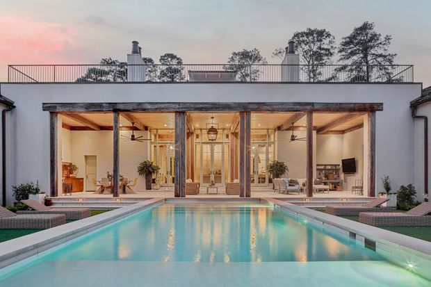 A symmetrical dusk view of a refined single-storey courtyard-style residence centered on a long rectangular swimming pool. The house has a white rendered facade, broad floor-to-ceiling glazed doors framed in dark timber, exposed timber roof beams and ceilings, two chimneys, and a rooftop terrace with slim metal balustrades. Lounge seating, potted plants, and lighting are visible through the glazing and around the pool, creating a warm connection between the interior and outdoor area.