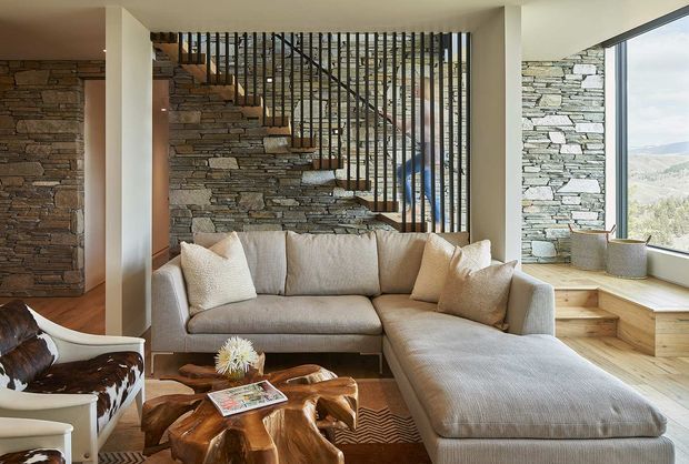 A warm contemporary living room with a large light-grey sectional sofa, sculptural live-edge wood coffee table, and cowhide accent chair. A stone-clad feature wall forms the backdrop to an open staircase with slender black vertical balustrades. Light timber flooring and broad corner glazing provide views toward a mountainous landscape.