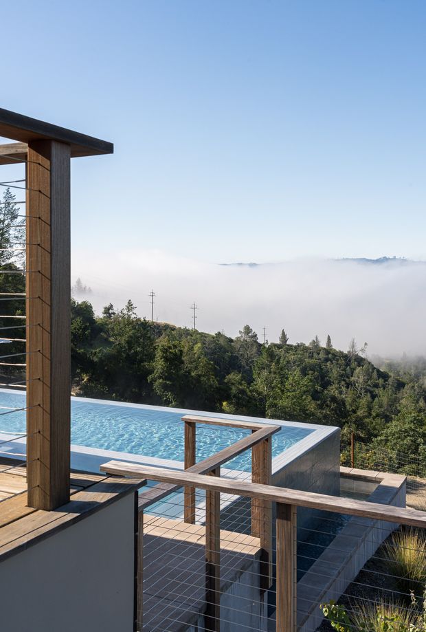 Exterior architectural photograph of a modern elevated terrace overlooking a wooded, mist-covered landscape. A rectangular swimming pool occupies the lower terrace, bordered by pale coping and dark paved surfaces. Timber posts and horizontal cable railings frame the deck and stairs, with a simple flat roof or canopy projecting at the left. The scene has clear blue skies, layered hills obscured by fog, and a restrained contemporary material palette.