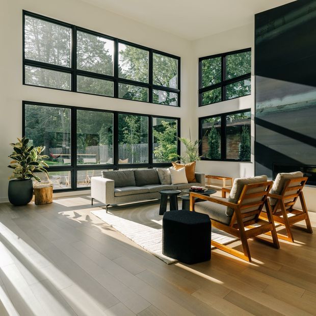 Bright contemporary living room with a double-height feel, expansive black-framed windows, pale timber flooring, a grey sofa, wooden lounge chairs, a low coffee table, and indoor plants. Strong natural light enters from the floor-to-ceiling glazing and overlooks a leafy garden. A dark vertical feature wall or fireplace element anchors the right side of the room.