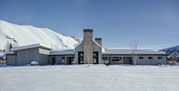 Wide exterior view of a low-slung contemporary mountain home set in deep snow, with a central steep-gabled entrance volume, tall stone-clad chimney, dark-framed glazing, and broad horizontal wings. Snow-covered alpine terrain and a clear blue sky surround the property.