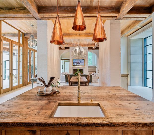 Warm, spacious open-plan kitchen and dining interior with a large rustic timber island in the foreground, integrated sink, three copper pendant lights, exposed timber ceiling beams, and pale textured masonry walls. Glazed timber doors line the left side, while the dining area and living space extend beyond the kitchen, creating a layered, welcoming composition.