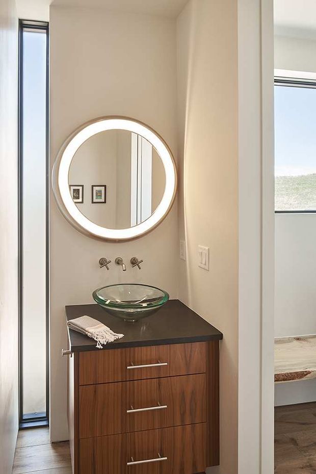 A compact powder-room vanity niche with a floating dark timber-grain cabinet, dark countertop, clear glass vessel basin, wall-mounted chrome tapware, and a large round illuminated mirror. Neutral walls and narrow vertical windows create a bright, refined interior; an adjacent bedroom is partially visible through the opening on the right.