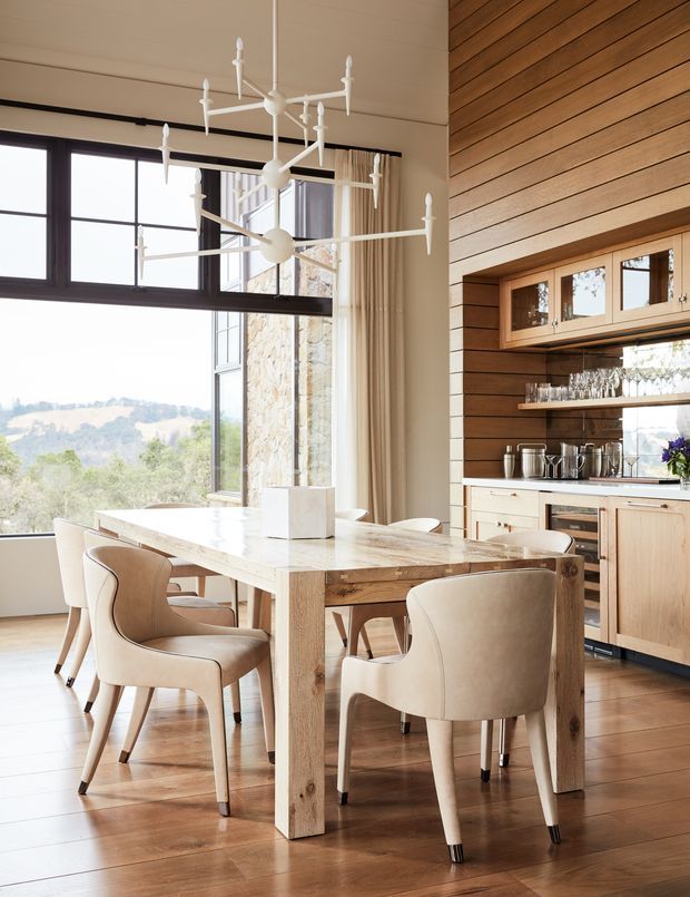 A bright, refined open-plan dining area with a large solid timber dining table and upholstered dining chairs. The room features warm timber flooring, a horizontal timber-lined feature wall, integrated timber cabinetry with glass-front storage, a stone-look benchtop, and expansive dark-framed glazing overlooking a green landscape. A sculptural white branching pendant light hangs above the table.
