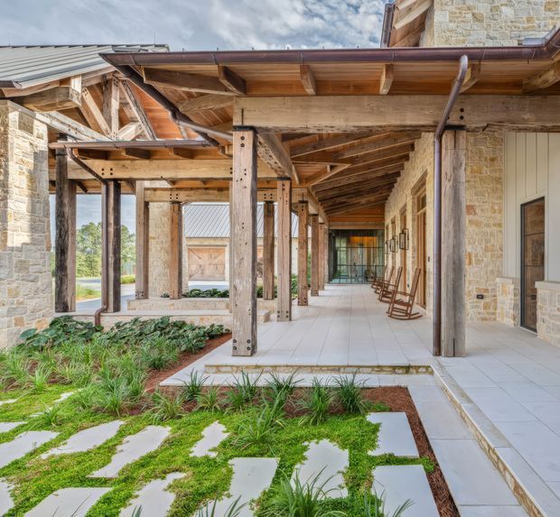 A covered exterior walkway and courtyard framed by heavy timber posts and exposed roof rafters. The building combines warm natural wood structure with pale rough-cut stone walls, light tiled paving, and a landscaped garden of grasses and groundcover. Rocking chairs line the veranda, while the open-sided passage connects multiple parts of the building. The image has a rustic, refined lodge-like character with strong indoor-outdoor circulation.