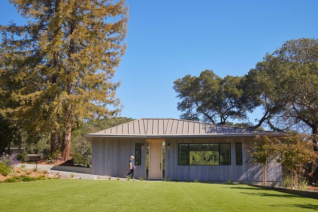 Single-storey contemporary house set within a landscaped suburban or rural garden, with a low-pitched standing-seam metal roof, vertically clad grey exterior walls, expansive dark-framed windows, and a recessed central entry. Mature trees and a broad lawn surround the building.