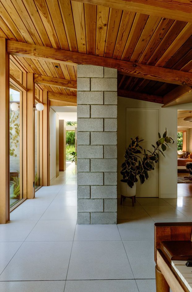 Interior view of a warm, timber-lined open-plan circulation and living area. The space features a slatted timber ceiling with substantial exposed timber beams, full-height timber-framed glazing along the left side, large pale square floor tiles, and a central exposed grey concrete-block structural partition. A doorway opens toward a furnished living area, while indoor plants and mid-century-style furniture add softness and character.