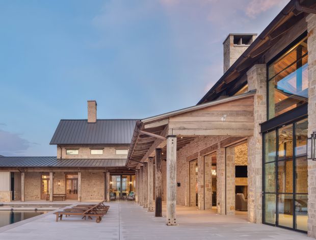 Exterior architectural photograph of a large stone-clad residence or lodge arranged around a paved courtyard and swimming pool. The building features pitched metal roofs with deep exposed timber eaves, heavy timber and stone columns, large black-framed glazed openings, and warm interior lighting visible through the glass. Pool loungers occupy the foreground, while the main emphasis is on the building facade and covered outdoor circulation space.