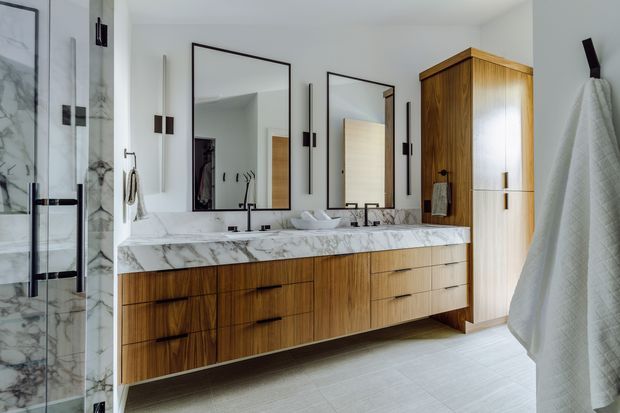 A bright, refined contemporary bathroom featuring a long double vanity with richly grained timber cabinetry, a white marble countertop and integrated backsplash, dark metal fixtures, and two tall rectangular mirrors. A marble-lined glass shower is visible at left, with a matching tall timber storage cabinet at right. The space has light flooring, white walls, and a clean, high-end finish.