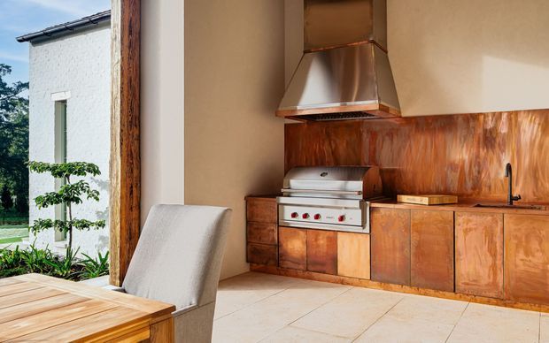 A partially open-plan indoor-outdoor kitchen with warm copper-toned timber cabinetry and backsplash, a built-in stainless steel barbecue and matching metal rangehood, and a dark tap over the benchtop. Pale large-format tiled flooring and neutral plaster walls create a warm, refined setting. A dining table and upholstered chair sit in the foreground, while a garden and adjacent white rendered building are visible through the large opening.