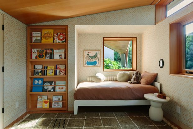 A compact, warmly lit bedroom or reading retreat with a built-in timber bookshelf filled with books, a low daybed upholstered in a soft neutral fabric, a small white sculptural side table, patterned wallpaper, timber ceiling, and timber-framed windows. A mirror reflects lush greenery and an exterior covered structure. The room has a cozy mid-century-inspired character with natural materials and abundant daylight.