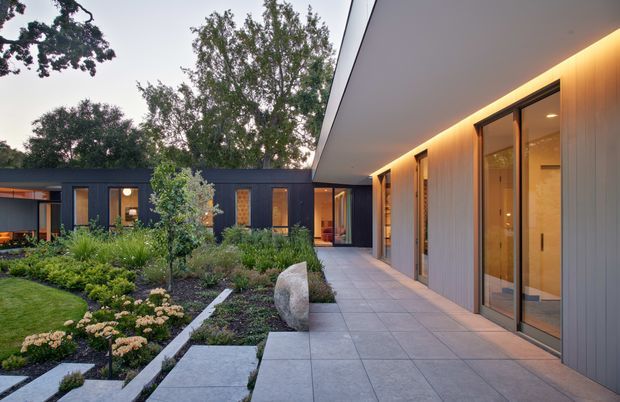 Contemporary single-storey residence arranged around a landscaped courtyard. The image looks along a broad paved walkway beside a long timber-clad wing with full-height glazed doors and warm concealed lighting, toward a darker-clad courtyard wing with recessed openings. Structured planting beds, ornamental grasses, clipped shrubs, stepping stones, and mature trees create a refined indoor-outdoor setting.