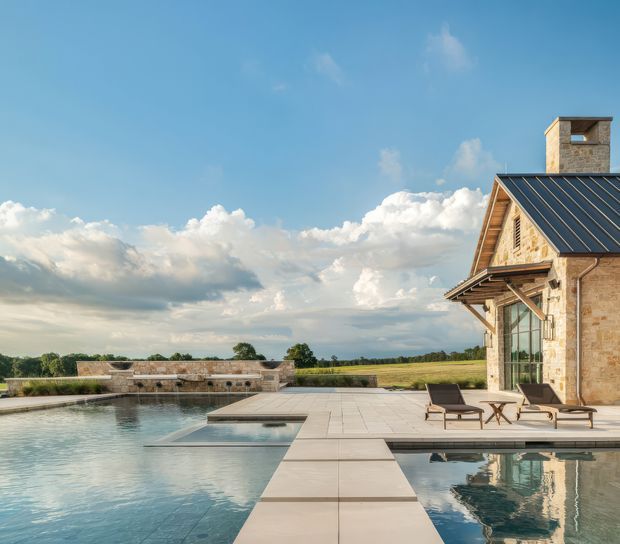 Exterior view of a substantial stone-clad house beside a large swimming pool, with broad paved terraces, lounge chairs, a dark standing-seam pitched roof, chimney, and expansive rural fields beyond. The scene is bright and open with a dramatic cloud-filled sky.