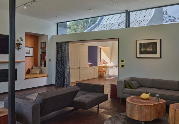 A contemporary living room viewed toward an adjoining dining area, with expansive clerestory glazing, a large open doorway, built-in window seat, dark upholstered seating, and a sculptural round timber coffee table. The room uses a restrained palette of white, charcoal, grey, and warm timber, with framed artwork and integrated shelving.