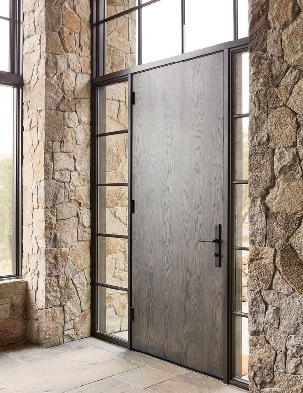 A tall timber entry door with dark metal hardware is set within a dramatic stone-clad wall, flanked by narrow black-framed glazed sidelights and tall windows. The entry has a pale stone floor and abundant natural light, creating a rustic yet refined architectural composition.