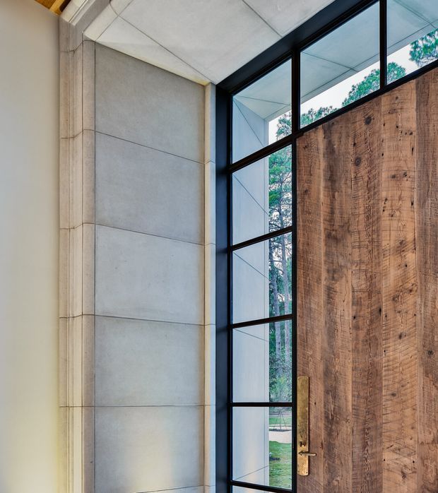 Close-up architectural view of a contemporary entry door beside a tall black-framed glazed sidelight. The door is clad in richly textured reclaimed-looking timber, contrasting with large smooth light-grey concrete or stone wall panels and a pale panelled ceiling. Trees and landscaped grounds are visible through the glazing, while warm concealed lighting highlights the entry surround.