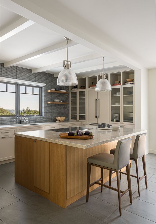 Bright, refined contemporary kitchen with a large wood-panelled island, pale stone benchtop, integrated cabinetry, glass-front display cupboards, open timber shelving, and two sculptural pendant lights. A dark grey tiled splashback frames the sink and window, while upholstered bar stools provide seating at the island. Exposed painted ceiling beams and large windows contribute to an airy, open feel.