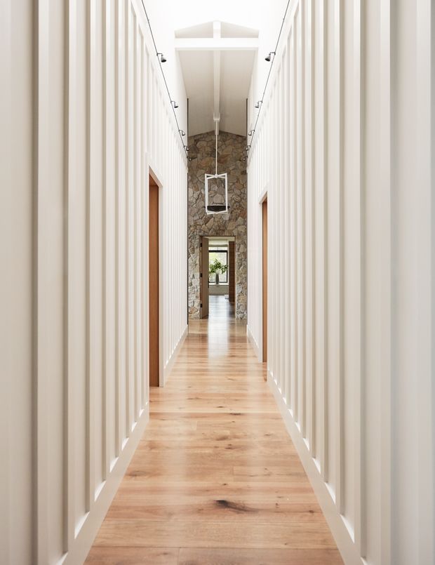 A long, narrow interior hallway with white vertical wall panelling, pale timber flooring, and a high pitched or vaulted ceiling. Slim black track lights run along both sides, while a large hanging pendant is suspended near the far end. The corridor terminates at a rustic natural-stone feature wall with an opening leading to another bright room. Warm timber doors and flooring contrast with the predominantly white, light-filled interior.