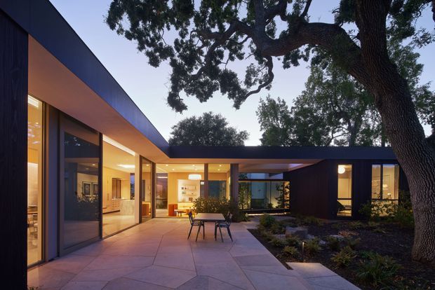 Contemporary single-storey courtyard house photographed at dusk, with low flat roof planes, extensive floor-to-ceiling glazing, dark cladding, warm interior lighting, and a broad paved terrace arranged around established trees and planted garden beds.