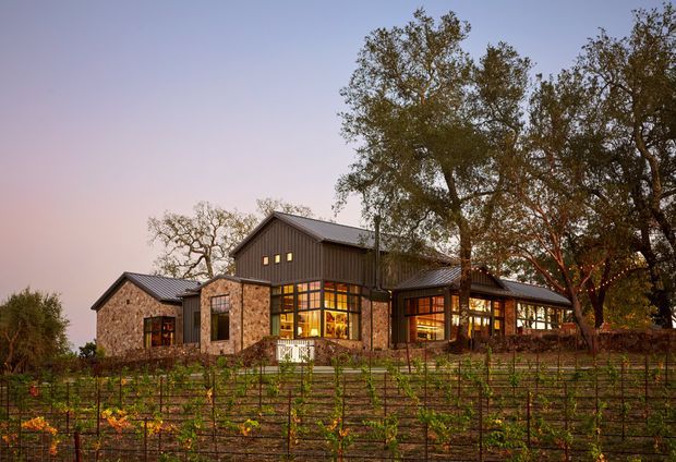 A large single-storey rural residence photographed at dusk, set behind a cultivated vineyard or garden. The home combines dark vertical cladding, extensive warm-lit glazing, natural stone walls, and multiple pitched roof forms. Mature trees surround the building, creating a secluded countryside setting.