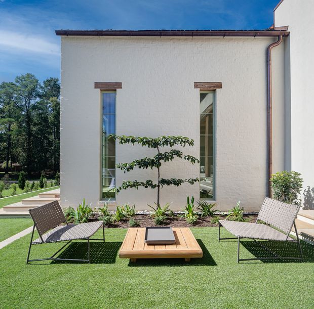 A sunny landscaped outdoor courtyard or terrace beside a white rendered building. The space features a low timber coffee table, two woven outdoor lounge chairs, artificial or manicured green turf, a planted garden bed with a small pruned tree and ornamental plants, and tall narrow windows with timber surrounds. The building has a simple pitched or shallow roof edge with exposed copper-toned rainwater goods, creating a refined contemporary garden setting.