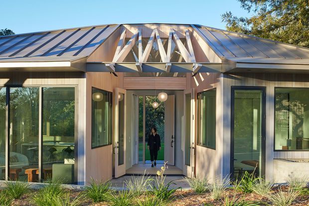 Front exterior view of a contemporary single-storey residence with a broad low-pitched metal roof and a recessed central entry. The facade combines warm vertical timber cladding, extensive glazed openings with dark frames, and an exposed timber truss feature above the entry. Native ornamental planting lines the foreground, while interior living areas are visible through the glazing.