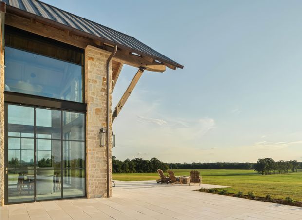 Exterior architectural view of a rural-style building with a steep standing-seam metal roof, exposed heavy timber roof structure and a pale natural-stone wall. Large black-framed glazed doors and windows open onto a broad paved terrace overlooking expansive green fields. Outdoor lounge chairs and a small table sit on the terrace, with warm late-day light and an open pastoral setting.