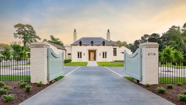 Formal front exterior view of a large symmetrical rendered residence behind open decorative gates. The home features a low hipped or mansard-like dark metal roof with dormer windows, twin chimneys, pale stucco walls, tall multi-pane windows, and a prominent timber entry door. A broad concrete driveway, masonry gate pillars, black metal fencing, clipped lawn, and landscaped planting create an elegant estate-style approach.