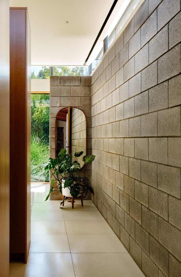 A narrow, light-filled interior passage with exposed grey concrete block walls, large pale floor tiles, a tall timber joinery element at left, and a rounded arched opening leading toward a garden-facing space. Indoor plants soften the corridor, while clerestory glazing and an open end bring in abundant natural light.